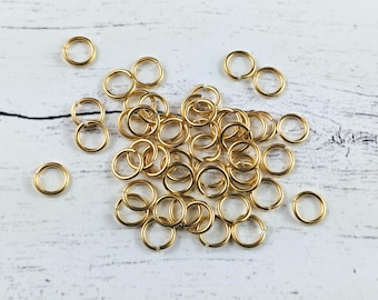 May include: A pile of gold jump rings, a type of jewelry finding, on a white wooden surface.