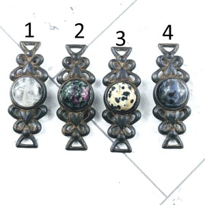 May include: Four antique drawer pulls with decorative metalwork and gemstone centers. The gemstones include clear, green with pink, speckled, and blue. Each pull is approximately 4 inches long.
