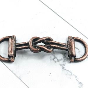 May include: A copper-toned metal buckle featuring a central knot design. The buckle has two D-shaped loops at each end, suitable for belts or accessories. The hardware is presented on a white and grey marble surface.