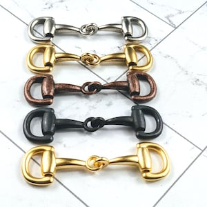 May include: An assortment of horse bit charms in silver, gold, bronze, and black. The bits are arranged in a row, showcasing their different metallic finishes. The charms have a classic equestrian design.