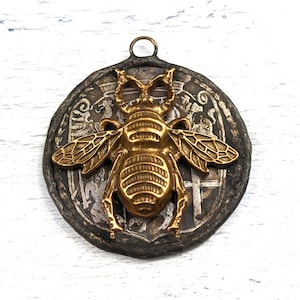 May include: A gold bee pendant with detailed wings and a textured body, set against a circular, silver-toned pendant with an intricate design.