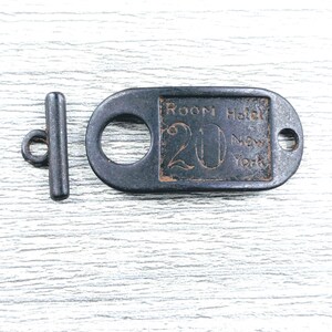 May include: A dark metal toggle clasp consisting of a T-bar and an oval plate. The plate features the embossed text Room Hotel 20 New York. The metal has a weathered, dark finish with visible texture and raised lettering on the rectangular centre.