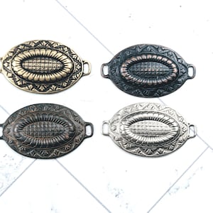 May include: Four oval-shaped metal embellishments in antique finishes: gold, black, bronze, and silver. Each has a detailed floral and geometric design with small loops on each end. The items are displayed on a white surface.
