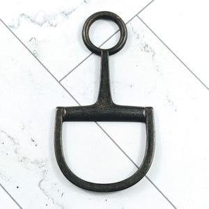 Puede incluir: Un bocado de caballo de metal oscuro, con una embocadura en forma de D y un anillo en la parte superior. El bocado tiene un aspecto envejecido y desgastado. El fondo es un patrón de mármol blanco y gris.