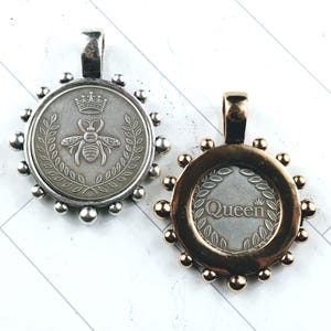 Könnte beinhalten: Zwei runde Anhänger mit einem Bienen- und Kronen-Design. Ein Anhänger ist silberfarben mit einer silbernen Öse, der andere goldfarben mit einer goldenen Öse. Der goldene Anhänger hat das Wort "Queen" in der Mitte.