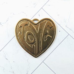 May include: A bronze-colored heart-shaped pendant with the word "LOVE" embossed in large, stylized letters. The pendant has a small loop at the top for hanging and is set against a white and gray patterned background.