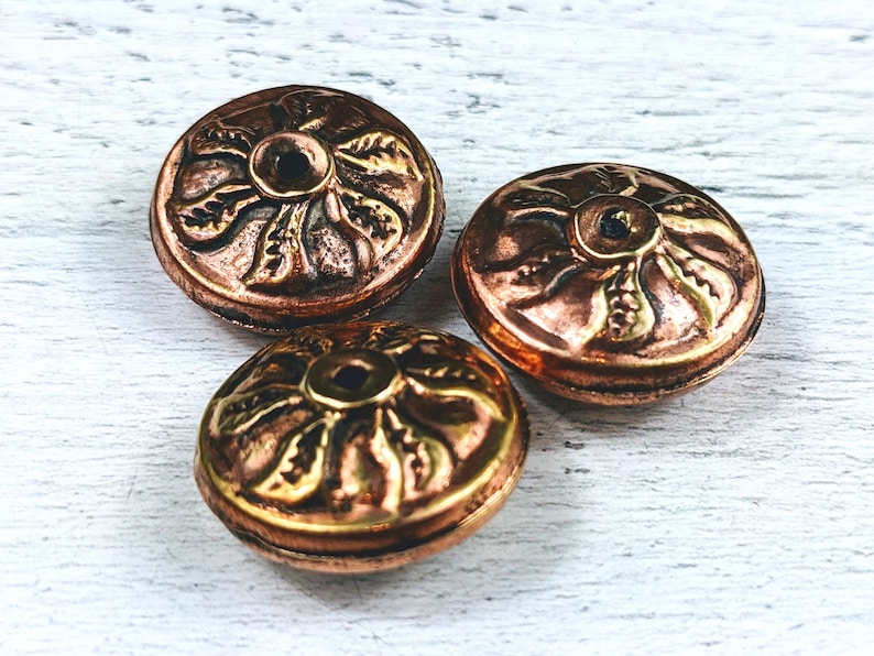May include: Three round, copper-colored metal beads with a decorative, floral design. The beads have a hole in the center and are approximately 1 inch in diameter.