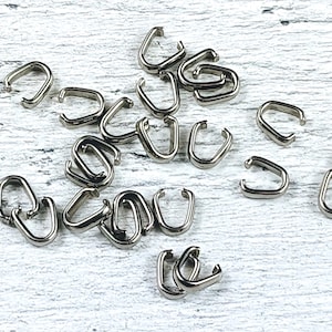 May include: A group of silver-toned metal jump rings, also known as split rings, are scattered on a white wooden surface. These rings are used for connecting jewelry components.