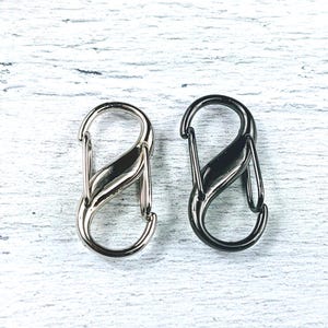 May include: Two metal S-shaped clasps, one silver and one black, on a white wooden surface.