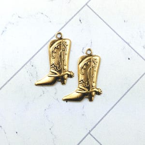May include: Two antique gold-colored cowboy boot charms. Each charm features intricate detailing, including decorative patterns and a spur. The charms have a loop at the top for easy attachment to jewelry or crafts.