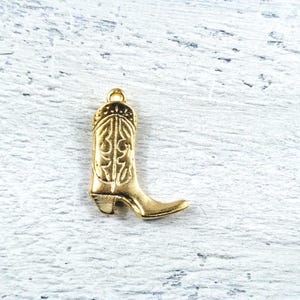 Könnte beinhalten: Ein goldfarbener Cowboy-Stiefel-Anhänger mit aufwendigen geprägten Details. Der Anhänger hat eine Schlaufe oben zur Befestigung an einer Halskette oder einem Armband. Der Stiefel hat eine spitze Spitze und einen detaillierten Absatz.