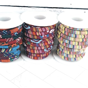 Puede incluir: Tres carretes de cuerda estampada de colores. La cuerda está enrollada alrededor de carretes blancos. Los patrones incluyen diseños geométricos en tonos de rojo, azul, naranja y amarillo. El fondo es una superficie de madera blanca.