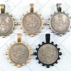 May include: Five coin-shaped pendants with a bee and crown design. The pendants have different colored frames: antique brass, gold, silver, and black. Each pendant has a loop at the top for a chain.