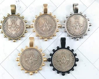 May include: Five coin-shaped pendants with a bee and crown design. The pendants have different colored frames: antique brass, gold, silver, and black. Each pendant has a loop at the top for a chain.