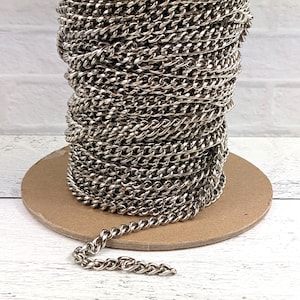 May include: A spool of silver-colored curb chain. The chain is tightly wound around a cardboard spool, with a length of chain extending out. The chain links are closely connected, creating a continuous, textured surface. Ideal for jewelry making and crafts.