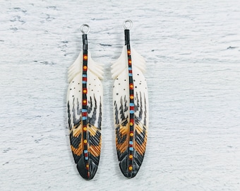 Puede incluir: Dos colgantes en forma de pluma de color crema con intrincados detalles tallados. Cada pluma presenta una punta negra, detalles en marrón y naranja, y una columna central con puntos rojos, azules y amarillos. Un lazo plateado en la parte superior para colgar.