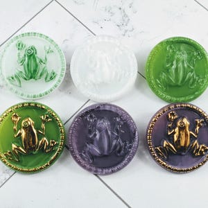 Könnte beinhalten: Sechs runde dekorative Knöpfe mit Froschmotiven. Die Knöpfe sind in verschiedenen Farben erhältlich, darunter Grün, Weiß und Lila. Einige Knöpfe haben goldfarbene Akzente. Das Froschdesign ist erhaben und detailliert.