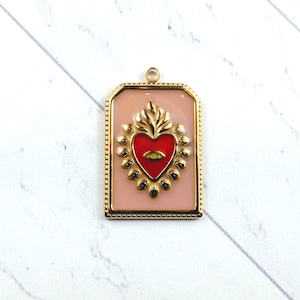 Puede incluir: Encanto de colgante de oro con fondo rosa y un corazón rojo con un ojo dorado en el centro. El corazón está rodeado por un diseño floral dorado.