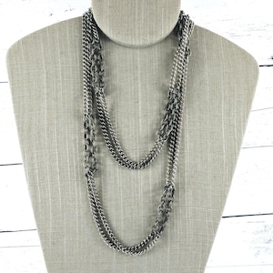 May include: A long, multi-strand necklace featuring a combination of polished silver-tone curb chains and larger, textured dark grey oval link chains. The layered design creates a cascading effect with varying chain sizes and metallic finishes.