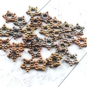 May include: A collection of star-shaped metal beads in various metallic finishes, including copper, bronze, and silver. Each bead features a central hole and small, rounded details around the edges. The beads are scattered on a white surface.