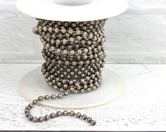 May include: A spool of silver-toned metal chain with round links. The chain is wound around a white spool and a section of the chain is hanging off the spool.
