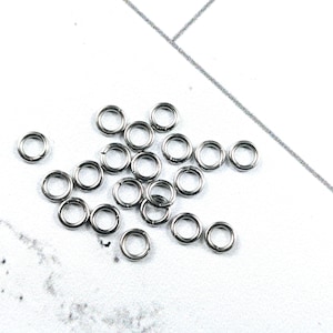 May include: A collection of small, silver-colored jump rings scattered on a white surface. The rings are circular and appear to be made of metal, likely for jewelry making or crafts. The background is a light, textured surface.