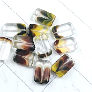 May include: A selection of clear, rectangular glass beads, each with a central hole. The beads display a marbled effect with brown, yellow, and green swirls. They are scattered on a white surface, creating a visually appealing arrangement.