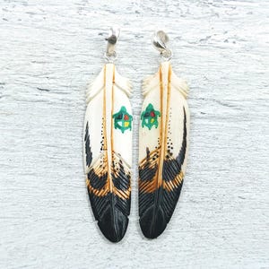 May include: Two feather-shaped pendants with silver-colored findings. The pendants are cream-colored with black, brown, and green accents. The feathers have a detailed design, with a small green and red emblem near the top.