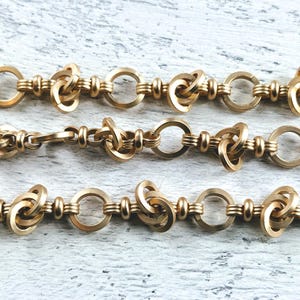 May include: A gold chain necklace with a unique knot design. The chain is made of multiple linked rings, each with a knot in the center. The chain is shown in three rows, with the knots facing the viewer.
