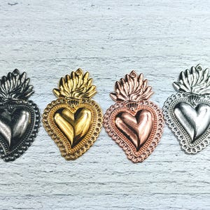 May include: Four ornate metal heart charms with a flame design. The charms are in gold, silver, rose gold, and black.