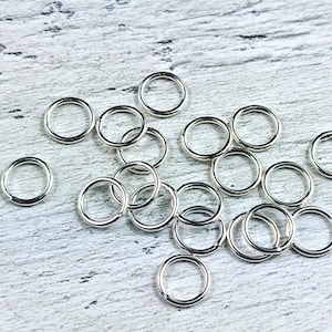 May include: A collection of silver jump rings, a type of jewelry finding, scattered on a white wooden surface.
