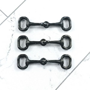 May include: Three dark gray metal horse bit charms. Each charm has a central ring connecting two oval-shaped ends. The charms are arranged in a vertical line against a white background with gray diagonal lines.