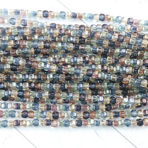 May include: Close-up of multiple strands of small, square beads in various colors. The beads are a mix of blue, green, brown, and clear, creating a textured and colorful appearance. Ideal for jewelry making or crafting.