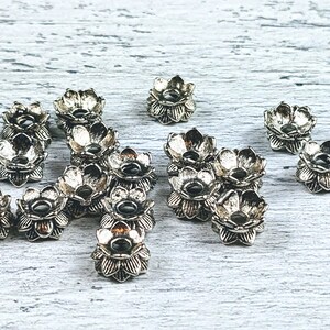 May include: A collection of silver metal flower-shaped beads. The beads have a raised design and a hole in the center for stringing.