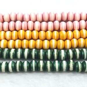 May include: Three strands of wooden beads with white stripes. The top strand is pink, the middle strand is yellow, and the bottom strand is green.
