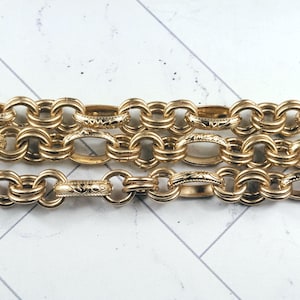 May include: Close-up of three strands of a gold-colored chain. The chain links are oval-shaped and connected, with intricate etched designs on some links. The chain is displayed against a white and gray surface.