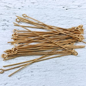 May include: A collection of gold-colored head pins with looped ends, scattered on a light-colored, textured surface. These pins are used for jewelry making and crafting. The pins are thin and have a consistent color.