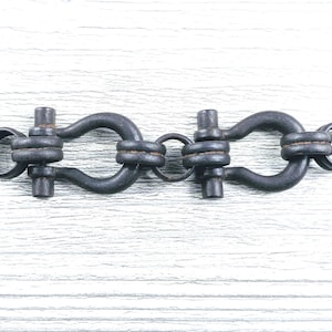 May include: A dark, matte-finished metal connector featuring two U-shaped shackle components linked by a central circular ring. The piece includes two cylindrical pins securing the shackles, with open circular loops at both ends of the assembly.