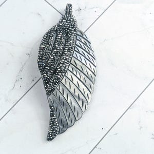 May include: A silver-toned leaf pendant with a textured design. The top portion is encrusted with small, sparkling black stones. The pendant has a loop at the top for a chain.