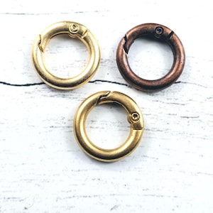 May include: Three metal spring rings in gold and bronze tones. The rings are circular with a hinged opening and a spring-loaded closure. The background is a white, textured surface.