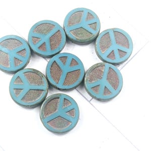 May include: A collection of eight round, turquoise-colored peace sign beads. Each bead features a raised peace symbol in a lighter shade of turquoise against a textured, brown background. The beads are arranged on a white surface.
