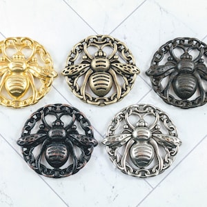 May include: Five decorative bee-shaped embellishments in various metallic finishes: gold, bronze, dark bronze, black, and silver. Each bee is centered within a circular, ornate frame. The bees have detailed wings and body segments.