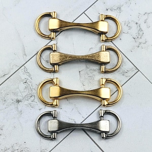 Peut inclure: Quatre connecteurs métalliques en forme d'anneau en or et argent. Les connecteurs ont une barre centrale avec des extrémités arrondies et deux anneaux en D de chaque côté. Le fond est une surface à motifs gris clair et blanc.