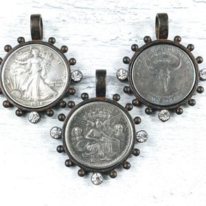 May include: Three silver coin pendants with a rustic, aged look. Each pendant features a different design: a walking liberty, a buffalo, and a winged angel. The pendants are surrounded by a circle of small, clear rhinestones.