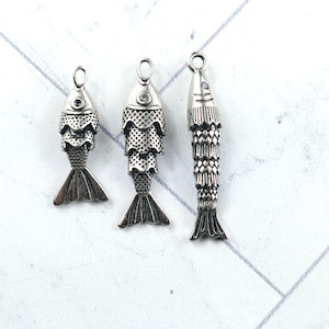 May include: Three silver-toned metal fish charms featuring detailed scale textures and small circular loops at the top for attachment. The charms vary in length and width, each displaying a distinct fish-shaped body with defined fins and tails.