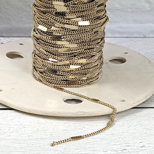 May include: A spool of gold-colored chain with a flat, rectangular link design. The chain is wound around a white ceramic spool.
