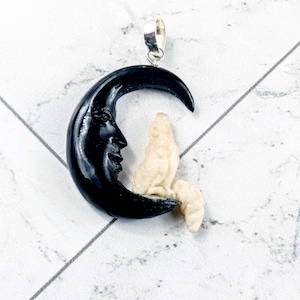 May include: A pendant featuring a black crescent moon with a carved face and a cream-colored wolf figure. The pendant has a silver-colored bail for attaching to a chain. The design is detailed and has a celestial theme.