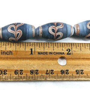 May include: Close-up of a row of blue and white glass beads with a red and white swirl design. The beads are approximately 1 inch long and are measured against a wooden ruler.