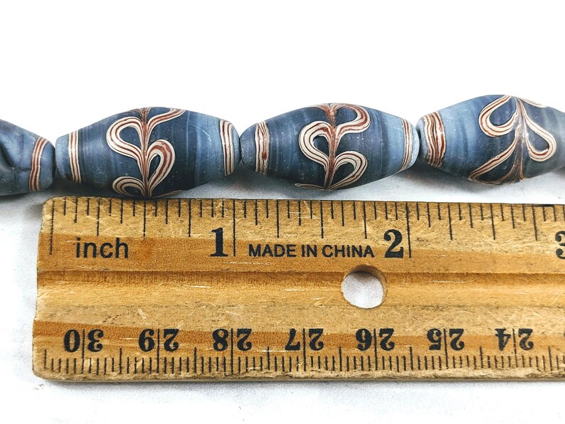 May include: Close-up of a row of blue and white glass beads with a red and white swirl design. The beads are approximately 1 inch long and are measured against a wooden ruler.