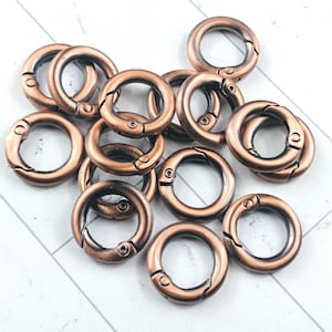 May include: A collection of copper-colored round spring snap clasps. These metal clasps are open and closed, showing their functionality. The clasps are scattered on a white and gray patterned surface.